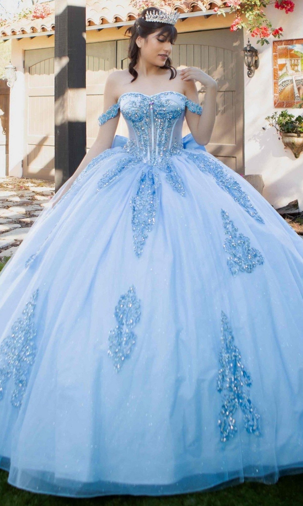 A woman wearing the Quinceanera Dress 8190J by Cinderella Couture in blue, featuring beaded details, stands outside in front of a house, looking to the side.